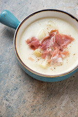 Potato soup served with torn prosciutto in a turquoise bowl, vertical shot on a light-brown granite background, middle closeup