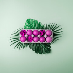 Purple Easter eggs in tray with flowers and tropical leaves on blue background. Top view. Creative Easter background.