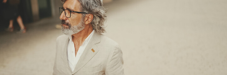 Panorama of mature businessman with beard in eyeglasses wearing gray jacket walks down the street...
