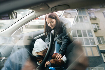 View through side window of car to young mother putting child safety seat with infant baby boy on front passenger seat with back to engine. Concept of safety driving and travel.