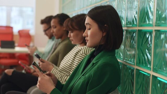 Tracking shot of diverse young business people, hiring, using phones in waiting room for networking, social media or communication at office. Group of employee workers wait in line with smartphone