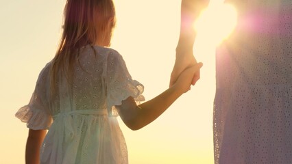 Happy family mother and daughter holding hands walking together at bright sun light sky closeup back view. Woman and girl child in elegant white dress going at natural sunlight sunny sunset sunrise