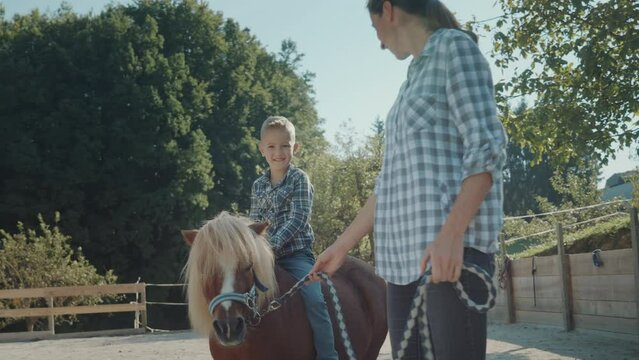Young boy riding brown pony with the instructor. Enjoying and riding ponies in the countryside on a sunny day. 