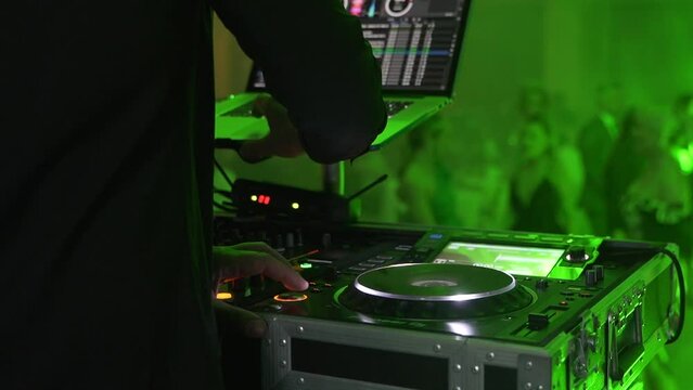 DJ hands mix at his desk, musical pieces for guests from the event. The guests dance in the background. Colored lights playing in the ballroom