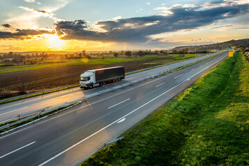 White transportation truck with trailer on an asphalt highway road in a rural landscape at sunset