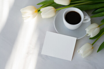 Obraz na płótnie Canvas Blank paper card mockup, coffee cup, saucer, white tulip flowers bouquet on neutral marble table background with abstract natural sun light shadows, good morning concept, postcard, invitation template
