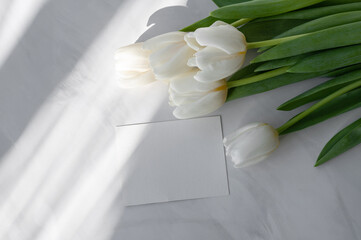 White tulip flowers bouquet and blank paper card mockup on neutral marble table with abstract natural sunlight shadows, aesthetic spring greeting postcard, business card or wedding invitation template