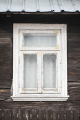 Wooden window frame. White peeling paint rustic architecture. Metal sheet roof. Abandoned home. Fence in front of a house. Lace curtains behind the glass. Rustic architecture.