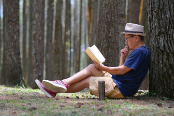 Man enjoying his leisure time sitting under a tree in the park, reading a book - obrazy, fototapety, plakaty