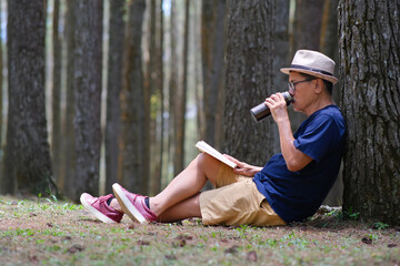 Man sitting under a tree in the pine forest, drinking cold drink from a tumbler. - obrazy, fototapety, plakaty