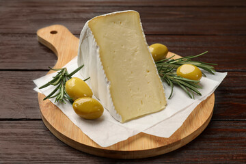 Board with piece of tasty camembert cheese, olives and rosemary on wooden table, closeup