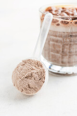 Whey protein powder in measuring spoon with glass of chocolate protein drink, milkshake smoothie on white table