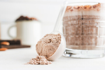 Whey protein powder in measuring spoon with glass of chocolate protein drink, milkshake smoothie on white table