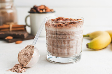 Glass with chocolate protein drink, milkshake smoothie on white table with bananas, protein powder...