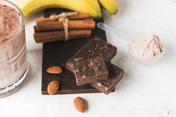 Pieces of chocolate protein bar, glass with protein drink, milkshake smoothie on white table with bananas, protein powder in measuring spoon, almond nuts and cinnamon sticks