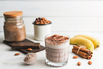 Glass with chocolate protein drink, milkshake smoothie on white table with bananas, protein powder in measuring spoon, protein bar, almond nuts and cinnamon sticks