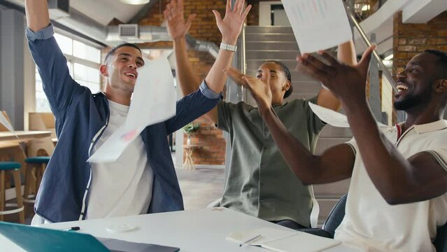Three young businessmen meeting in modern office sitting at table with laptop celebrating throwing documents into the air - shot in slow motion