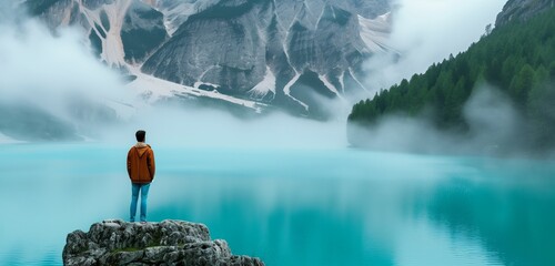 Side view of man standing in view of turquoise lake with foggy mountains
