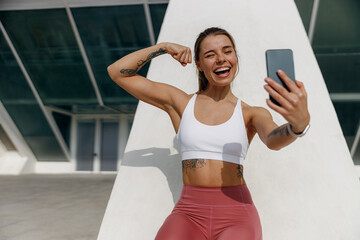 Sporty smiling young woman after running making selfie outdoors and showing biceps