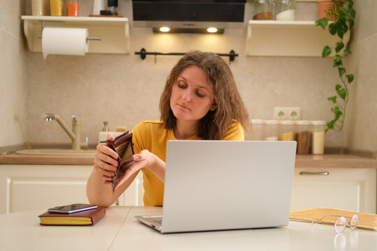 A woman with a laptop holds an empty wallet without money while sitting at a table in a home kitchen. An adult female businesswoman works from home, a remote office