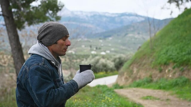Young man in denim jacket relaxing alone in mountains, drinking hot coffee from thermos mug, enjoys happy weekend trekking in nature. Travel lifestyle concept. National park Sierra de Cazorla in Spain
