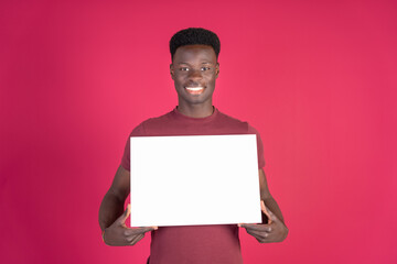 Portrait of cheerful young black man in red t-shirt holding and showing blank empty horizontal sign standing isolated on carmin red studio background, copy space. Concept space for advertising.