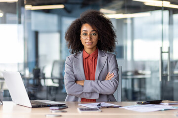 A professional businesswoman standing confidently at her workspace with her arms crossed. She...