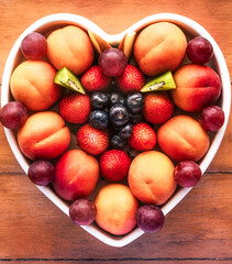 Close-up view from above of a white heart-shaped plate full of red grapes, apricots, strawberry and blueberries. Wooden background