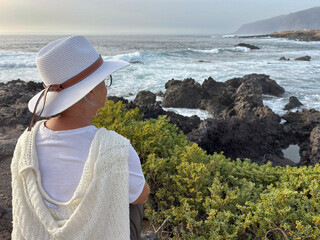 Rear view of senior woman sitting on a rocky beach watching ocean waves crashing with white foam....