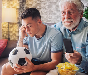 Young boy and senior bearded grandfather soccer fans watching a football game on tv sitting on a...