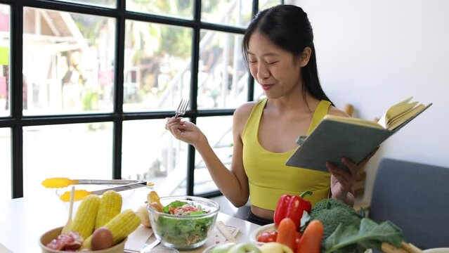 Young Asian woman at home eating vegetable salad and reading health care book.