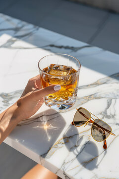 High Fashion woman's hand holding a rocks glass cocktail on a marble table with yellow and gold sunglasses.