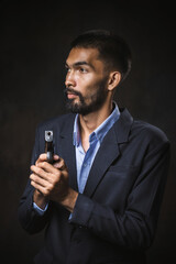 A bearded young man wearing a suit holding a pistol. Male mafia assassin or intelligence agent holds a gun on black background.