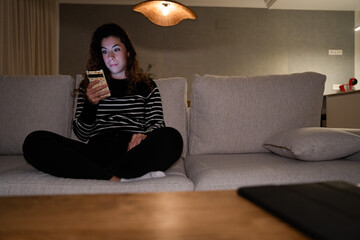 A woman is sitting on a couch and looking at her phone surprised. She is wearing a black and white striped shirt