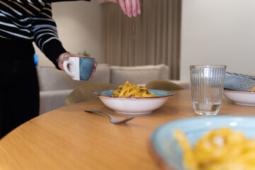 A person is pouring cheese on a plate of pasta. The table is set with a bowl of pasta, a cup, a fork, and a spoon