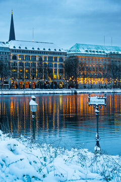 Blick auf das mit Schnee bedeckte Schild Lombardbrücke, im Hintergrund Weihnachtsbeleuchtung und beleuchtete Hausfassaden im Winter, die sich im Wasser spiegeln, vertikal, horizontal