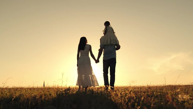 Silhouettes of woman with husband carrying daughter on shoulders walking hand in hand. Woman looks at daughter sitting on shoulder of father on field. Woman with boyfriend and daughter on shoulders