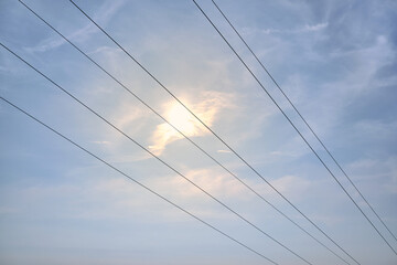 Diagonal hanging high-voltage transmission power lines against blue sky with clouds and shining...