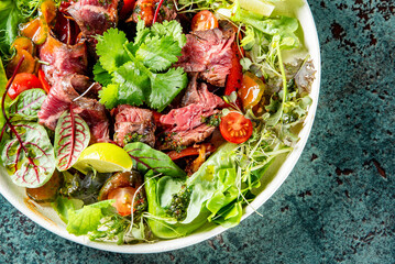 Vibrant steak salad served in white bowl. Features juicy steak slices, fresh greens including spinach and lettuce, garnished with coriander leaves and cherry tomatoes. Presented on a textured surface