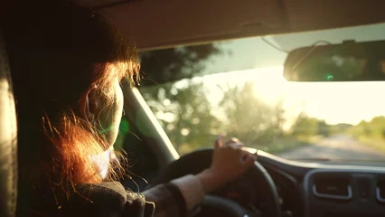 Fotobehang Redhead woman driver driving car at sunlight countryside rural summer landscape road trip closeup back view. Modern female riding automobile holding steering wheel enjoy automobile movement on path © SUPER FOX