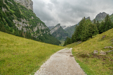 Fototapeta na wymiar Hikinig trail towards Seealpsee, an alpine lake in Appenzell Alps in Switzerland. Easey walking path to an alpine lake, pine trees on the side of the trail, alpine lake between steep mountains. Swiss.