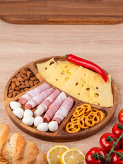 Top view of a round wooden plate with different snacks: sausages, cheese, peppers, standing on a wooden table.