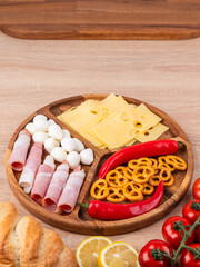 Top view of a round wooden plate with different snacks: sausages, cheese, peppers, standing on a wooden table.