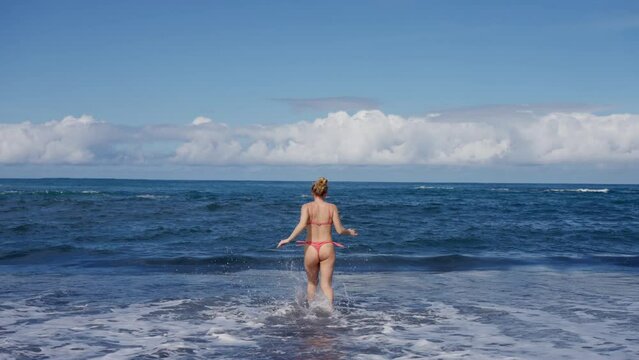 Girl in a swimsuit runs along the beach and looks at the water and the horizon. Her gesture shows admiration for the vast ocean, sky and clouds above. view from the back, slow motion