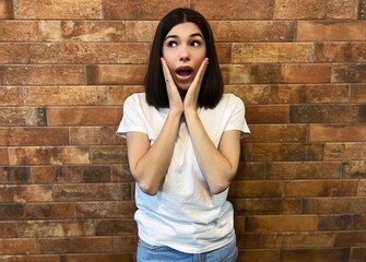 young brunette woman surprised and shocked, expressing surprise. Brunette in a white T-shirt posing in surprise against the background of a brick wall. 