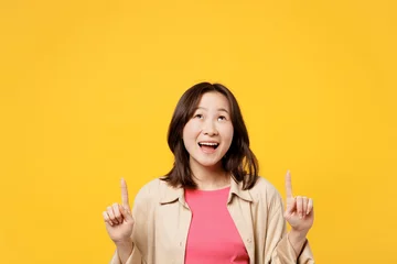 Fotobehang Young happy woman of Asian ethnicity she wear pink t-shirt beige shirt pastel casual clothes point index finger overhead on area isolated on plain yellow background studio portrait Lifestyle portrait © ViDi Studio