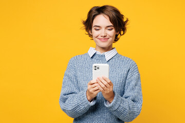Young smiling happy woman she wearing grey knitted sweater shirt casual clothes hold in hand use mobile cell phone chat online isolated on plain yellow background studio portrait. Lifestyle concept.