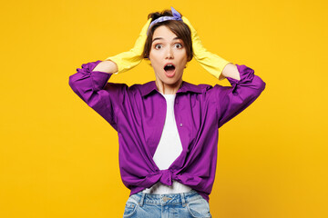 Young surprised shocked astonished woman wear purple shirt rubber gloves while doing housework tidy up look camera hold head isolated on plain yellow background studio portrait. Housekeeping concept.