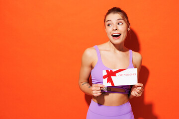 Young fitness trainer sporty woman sportsman wear purple top clothes in home gym hold gift coupon voucher card for store look aside isolated on plain orange background. Workout sport fit abs concept.