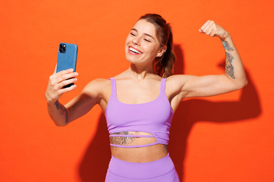 Young fun fitness trainer woman sportsman wear top shorts purple clothes in home gym do selfie shot on mobile cell phone show biceps isolated on plain orange background. Workout sport fit abs concept.